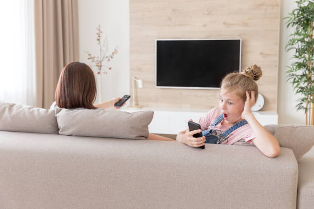 istock Girl sitting on couch and watching something interesting in mobile phone while mother watching tv. surprise concept 1170053295