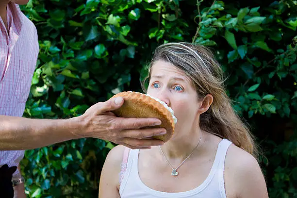 Head and shoulders image of a woman looking frightened at the hand of her husband which is holding a custard pie.