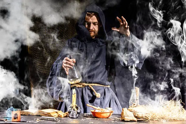 The medieval alchemist make magic ritual at the table in his smoke laboratory. Halloween concept background.