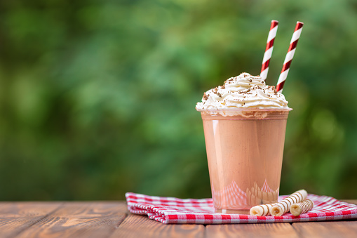 chocolate milkshake in disposable plastic cup with whipped cream and wafer rolls on wooden table outdoors