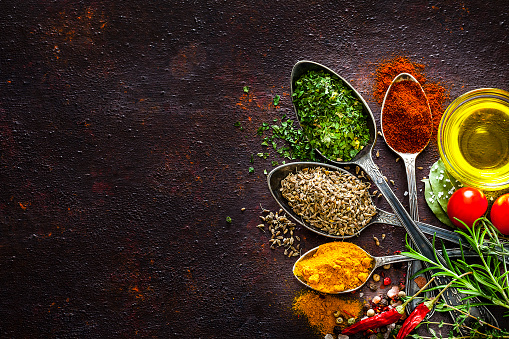 Top view of four vintage spoons filled with multi colored spices and herbs placed at the right border of a rustic brown background leaving useful copy space for text and/or logo at the center left. Spices and herb included are turmeric, bay leaf, curry powder, peppercorns, salt, chili pepper, rosemary and olive oil. Low key DSRL studio photo taken with Canon EOS 5D Mk II and Canon EF 100mm f/2.8L Macro IS USM.