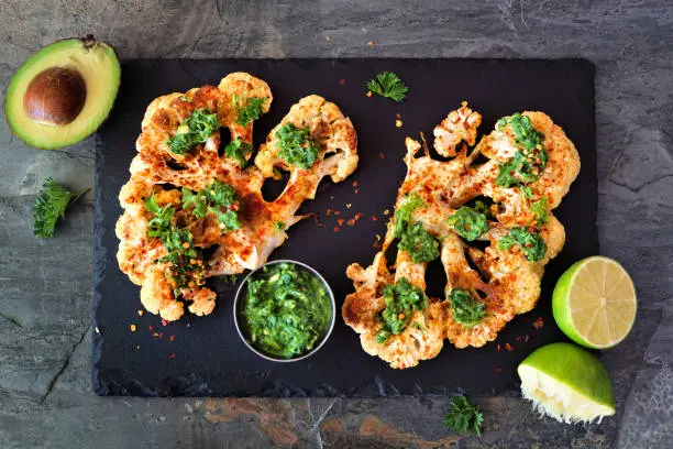 Cauliflower steaks with a cilantro lime sauce. Above view on a slate platter. Healthy eating, plant based meat substitute concept.