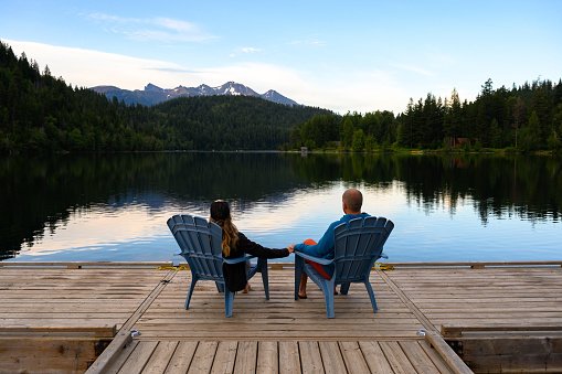 Rear view of couple relaxing on Adirondack chairs at lake. Male and female are spending leisure time together. They are on pier.