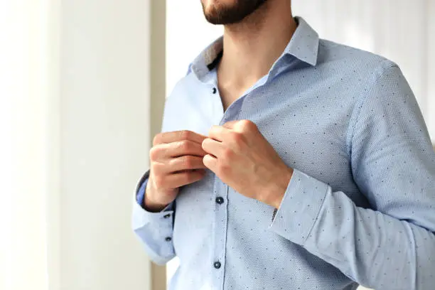 Photo of Man buttons sleeves on blue linen dress shirt in bedroom at slight angle.