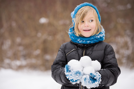 Portrait of adorable little kid boy with long blond hair playing with snowballs outdoors. Child with blue scarf and hat walking and having fun on a windy winter day.