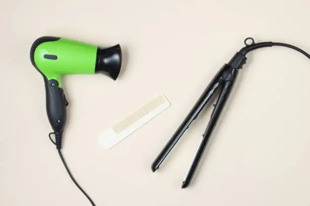 Photo of Minimalistic beauty and fashion still life. Hair dryer, comb and hair straightener on beige background. Top view, flat lay