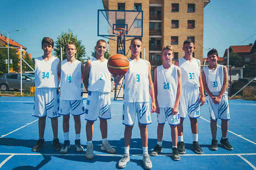 Portrait of young guy playing basketball on an outdoor game court, girl is out of focus. Handsome teenager smiling looking at camera with ball in hands in sunset light. Youth, active healthy lifestyle