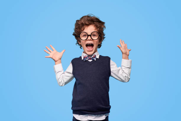 Excited schoolboy screaming and gesturing with hands Little genius in nerdy glasses and school uniform yelling in excitement and holding hands up against blue background nerd kid stock pictures, royalty-free photos & images