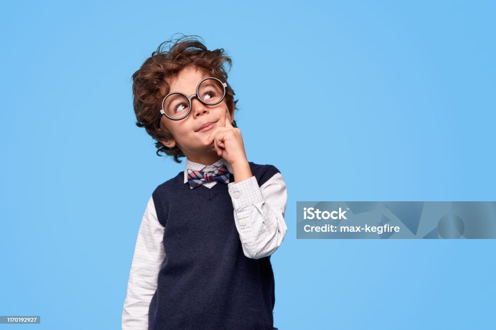 Thoughtful little genius looking up Smart wunderkind in nerdy glasses and school uniform touching cheek and looking up while thinking against blue background Child Stock Photo