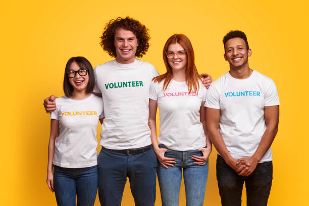 Multiethnic volunteers smiling for camera Group of diverse young people in volunteer T-shirts cheerfully smiling and looking at camera against vivid yellow background altruism photos stock pictures, royalty-free photos & images