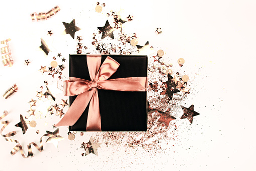 Black gift box with rose gold bow on white background with glitter. Holiday concept.