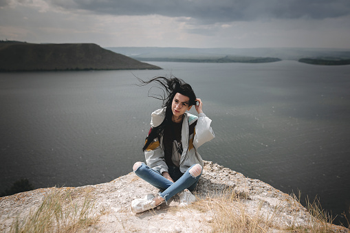 Stylish hipster girl with windy hair sitting on top of rock mountain with beautiful view on river. Young tourist woman relaxing on cliff. Travel and wanderlust. Copy space