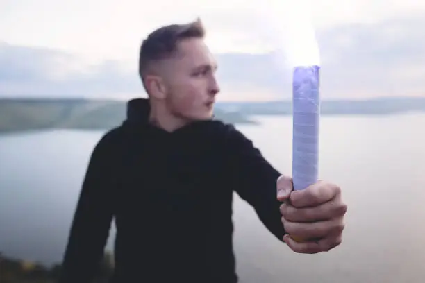 Photo of Ultras hooligan holding blue flare torch in hand, standing on top of rock mountain in evening with amazing view on river. Atmospheric moment. Copy space