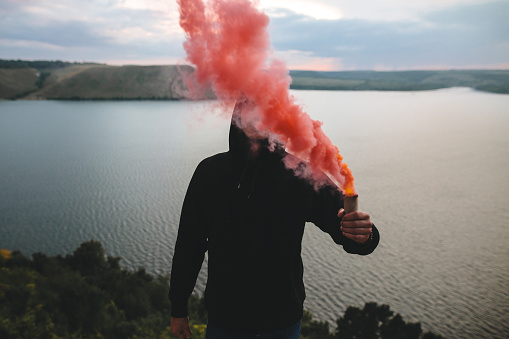 Ultras hooligan holding red smoke bomb in hand, standing on top of rock mountain with amazing view on river. Atmospheric moment. Traveler guy with smoke. Copy space