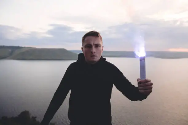 Photo of Ultras hooligan holding blue flare torch in hand, standing on top of rock mountain in evening with amazing view on river. Atmospheric moment. Copy space