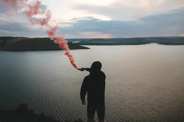 Photo of Ultras hooligan holding red smoke bomb in hand, standing on top of rock mountain with amazing sunset view on river. Atmospheric moment. Traveler guy with smoke. Copy space