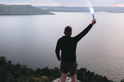 Ultras hooligan holding blue flare torch in hand, standing on top of rock mountain in evening with amazing view on river. Atmospheric moment. Copy space