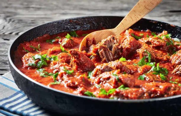 close-up of spicy Beef Stew in tomato sauce with spices and herbs in a skillet on a rustic wooden table, west African cuisine, horizontal view from above,