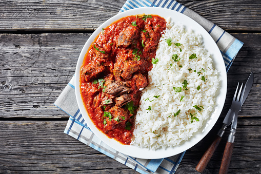 close-up of Beef Stew in tomato sauce with spices and herbs served on a plate with rice on an old wooden table, west African cuisine, horizontal view from above, flatlay, empty space