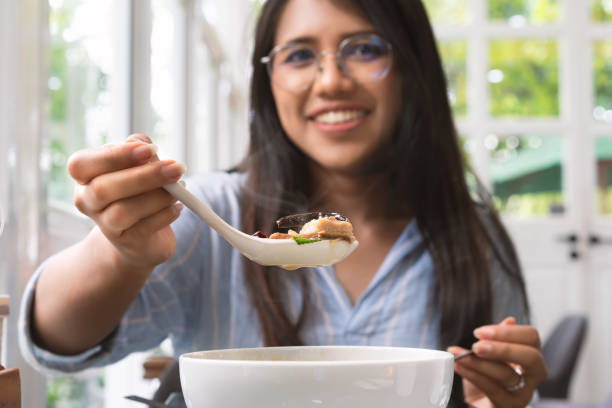 Asian woman holding up a spoon of Thai food to the camera in a trendy cafe Second person point of view of Asian woman holding up soup spoon of hot beef spicy Thai broth - Young diverse female serving hot steaming food to a guest diner - meal, cuisine and review concept hot vietnamese women pictures stock pictures, royalty-free photos & images
