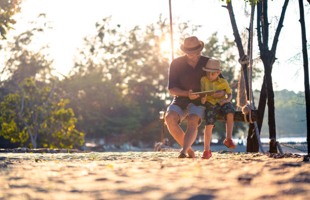 отец и сын с помощью цифрового планшета сидя качели - child beach digital tablet outdoors стоковые фото и изображения