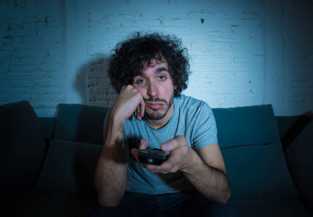 Young bored man on couch using TV remote control zapping for another movie or show late at night. Looking disinterested and sleepless. In entertainment People insomnia and Sedentary lifestyle concept. stock photo