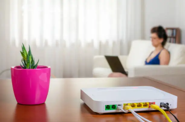 Modem router on a table in a living room. A woman using a laptop while sitting on the sofa is in background. Selective focus.