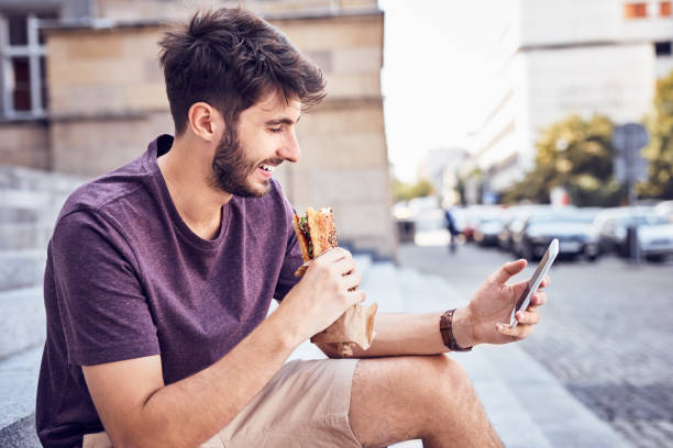 Young man eating lunch and using phone outdoors in the city