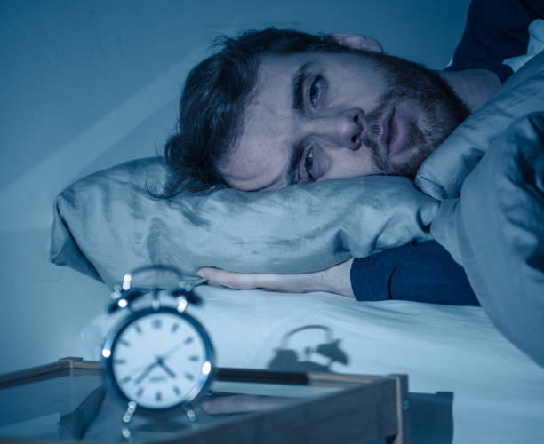 Sleepless and desperate young caucasian man awake at night not able to sleep, feeling frustrated and worried looking at clock suffering from insomnia in stress and sleeping disorder concept. stock photo