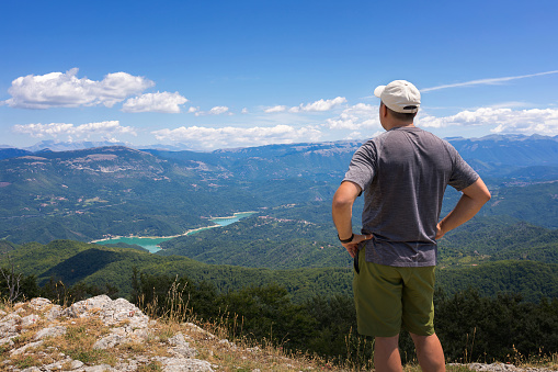 The man looks at the landscape on the horizon, from the top of a mountain. The hiker scrutinizes the mountain landscape and the lake below. On a summer day with cloudy skies.
