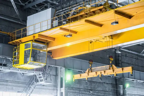 Photo of Yellow overhead crane with linear traverse and hooks in engineering plant shop. Cabin of crane operator and jib crab trolley. Landing  staircase.