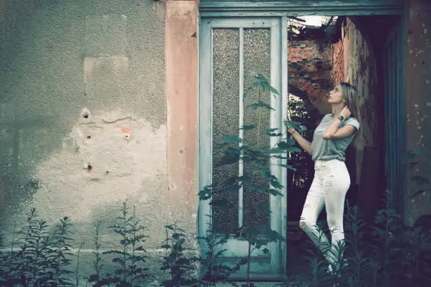 Sexy woman standing in the door to a dilapidated abandoned building daydreaming looking up into the air in a vintage style faded aged portrait