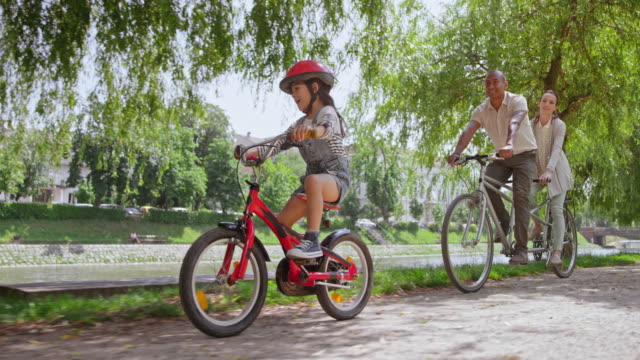 SLO MO TS Couple riding a tandem bike and their young daughter is riding beside them through the park