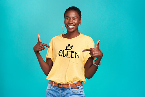 Studio shot of a confident young woman wearing a t shirt with “queen” on it against a turquoise background