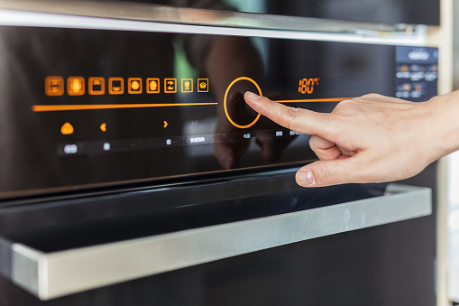 cropped shot of female hand choose mode with knob switch on control panel of modern built in oven, baking at home kitchen with stylish interior