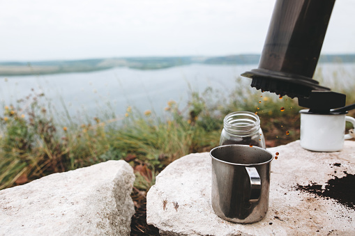 Coffee drops from aeropress on metal mug on cliff at lake, brewing alternative coffee at camping. Making hot drink at picnic outdoors. Trekking and hiking in mountains