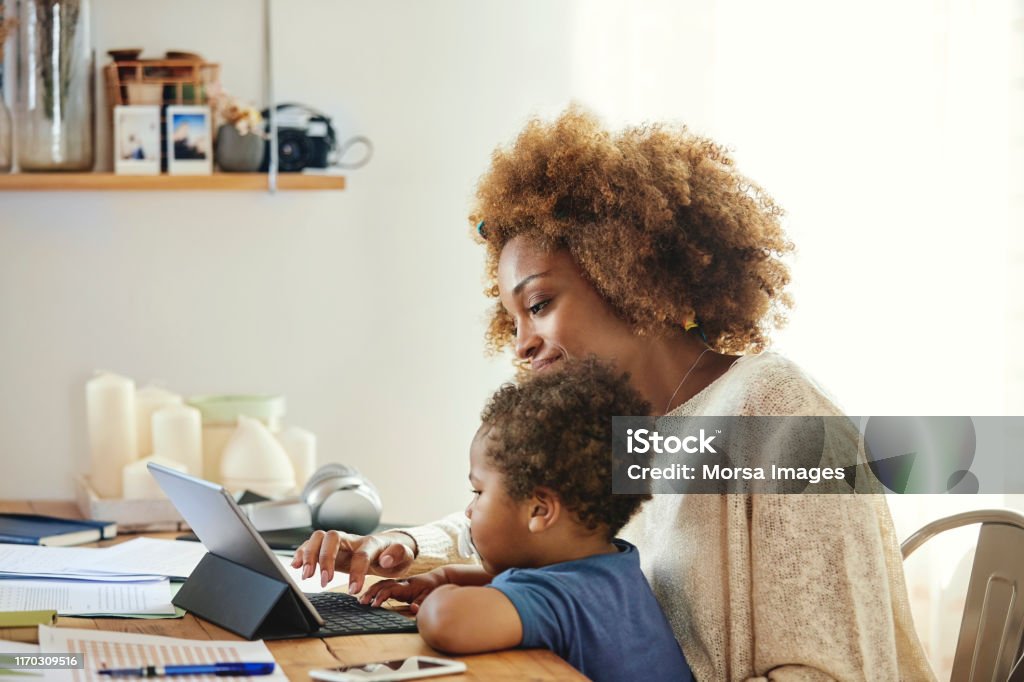 Smiling woman with son holding digital tablet Smiling mother with son using tablet computer. Woman sitting with boy at table in kitchen. They are wearing casuals. Baby - Human Age Stock Photo