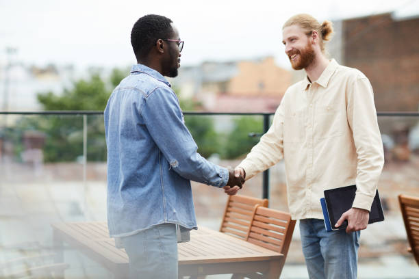 Business handshake before meeting Two young multiethnic colleagues standing shaking hands and smiling to each other before meeting in outdoor cafe casual handshake stock pictures, royalty-free photos & images