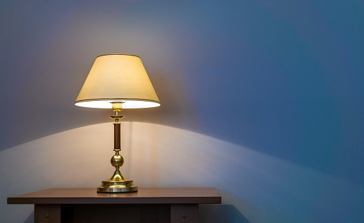 Classic table lamp with turned on bulb under lampshade is against wall with blue wallpaper. Copy space