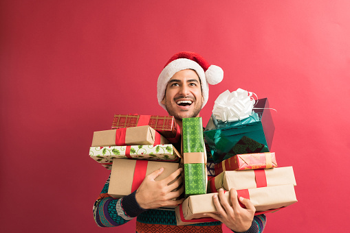 Cheerful young male looking up while carrying Xmas gifts against red background