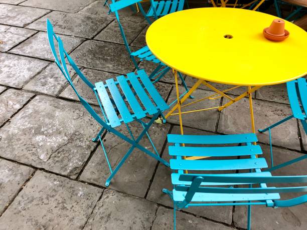 Image of colourfully painted, circular round metal and wooden garden patio tables and chairs in bright colours with rainbow paint, yellow table and turquoise blue chairs Stock photo showing coloured painted, small circular round metal and wooden garden patio table and chairs in bright colours, yellow and turquoise blue. outdoor dining photos stock pictures, royalty-free photos & images
