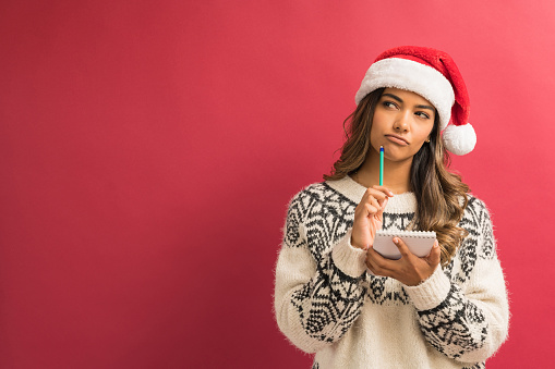 Young female Hispanic brunette looking away while holding notepad and pencil thinking about Christmas list against plain background