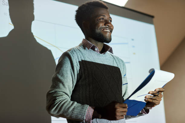 istock Smiling successful Black student with beard standing against projection screen in university room and reading report in front of audience 1170371803