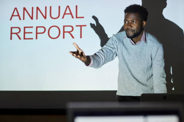 istock Content confident young Afro-American speaker standing at projection screen with ANNUAL REPORT slide and answering questions of colleagues at meeting 1170371879