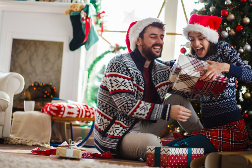 Copy space shot of jolly young couple sitting on the floor in their living room under Christmas and having excited looks on their faces after opening gifts.