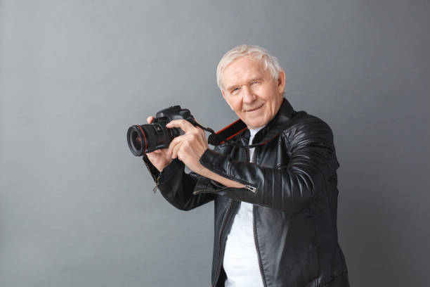 istock Senior man in leather jacket standing isolated on gray holding camera side view looking camera cheerful 1170400562