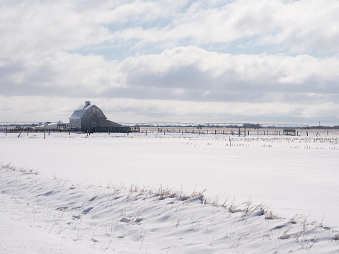 Old wooden barn with a snowy field in the foreground. Cloudy skies are above.