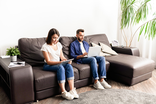 Concentrated young couple in casual clothing sitting on comfortable sofa and using devices while working as freelancers at home