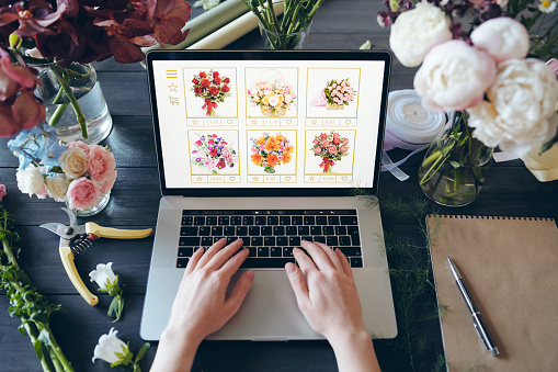 Above view of unrecognizable florist standing at desk with beautiful flowers and hand tools and typing on laptop keyboard while making online order of flowers for own shop