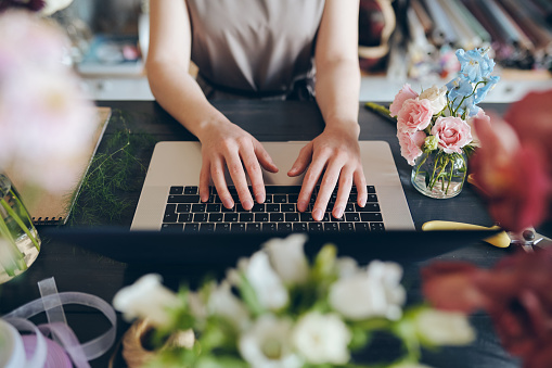 Close-up of unrecognizable female florist standing at desk and typing on laptop while working with online orders